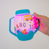 Hand holding a blue Glo Pals sensory jar glowing pink, decorated with ABC letters, numbers and a school bus