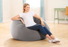 A woman sits comfortably on a large, round, gray bean bag chair in a bright, modern room with light wood floors and soft blue curtains.