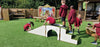 Children in maroon uniforms play on a wooden outdoor playground structure with a white arched bridge and steps on artificial grass, with a wooden fence and playhouse in the background.