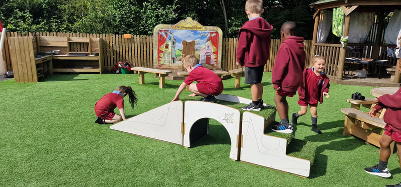 Children in maroon uniforms play on a wooden outdoor playground structure with a white arched bridge and steps on artificial grass, with a wooden fence and playhouse in the background.