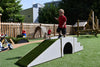 A young child in a red shirt and dark shorts stands on a white and green artificial turf play ramp in an outdoor children's playground with wooden fencing, other play equipment, and trees in the background.