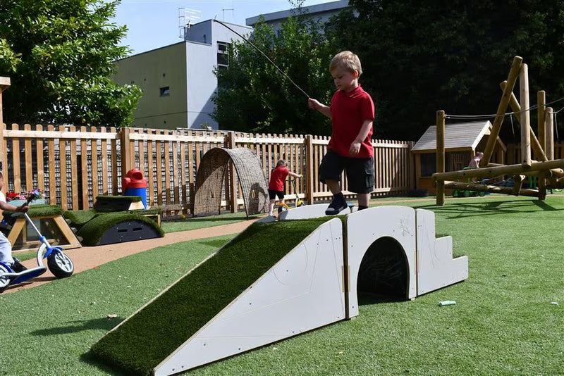 A young child in a red shirt and dark shorts stands on a white and green artificial turf play ramp in an outdoor children's playground with wooden fencing, other play equipment, and trees in the background.