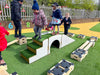 Children playing on an outdoor wooden obstacle course with grass-covered steps and bridges on artificial turf, with a white fence and trees in the background.