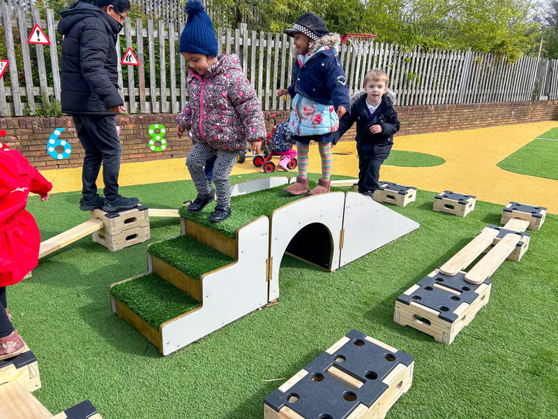Children playing on an outdoor wooden obstacle course with grass-covered steps and bridges on artificial turf, with a white fence and trees in the background.
