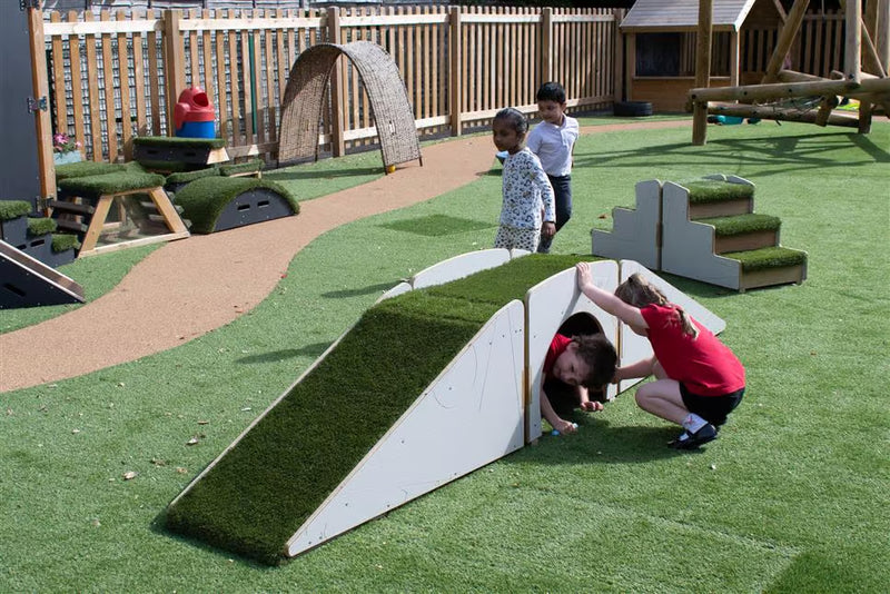 A children's outdoor play area with artificial grass surfaces, a wooden fence, and play equipment. In the foreground, a child in a red shirt crouches beside a white ramp covered in green turf, while another child peers out from under it.