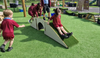 Children in red uniforms playing on a small outdoor playground structure with a slide and tunnel on artificial green turf.