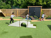 Outdoor play area with children using a white wooden arch bridge and ramp on green artificial turf, with a wooden fence, black playshed, and trees in the background.