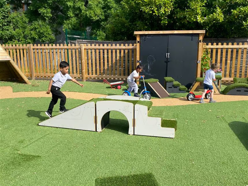 Outdoor play area with children using a white wooden arch bridge and ramp on green artificial turf, with a wooden fence, black playshed, and trees in the background.