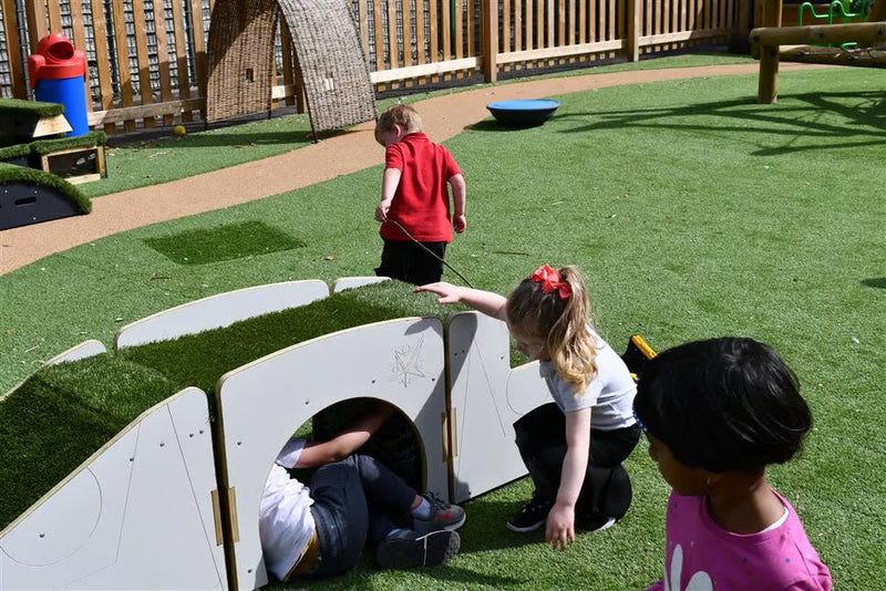 Children playing outdoors on artificial green turf near a white modular play structure with grass-covered roofs. One child is crawling through a round opening, another crouches beside it, and a third stands nearby.