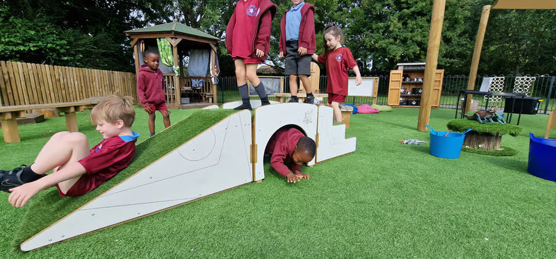 Children in maroon uniforms playing on a wooden outdoor play structure with a grass-covered slope and tunnel, set on artificial green turf in a schoolyard.