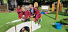 Four young children in maroon school uniforms play on a wooden cat tunnel and climbing structures on an outdoor artificial grass play area at a school.