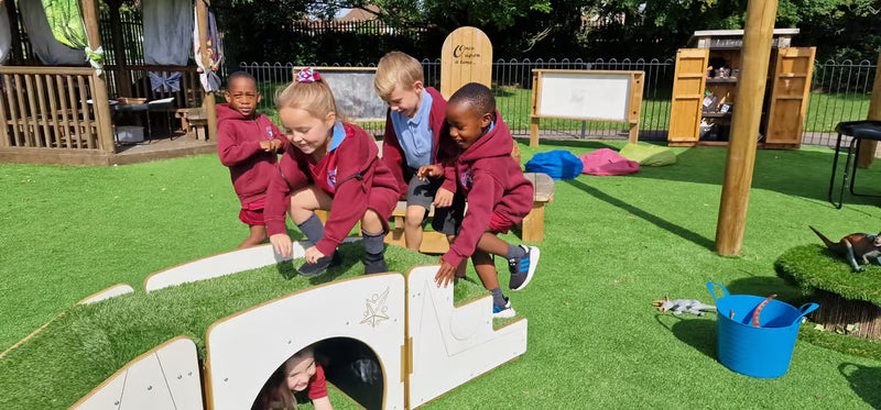 Four young children in maroon school uniforms play on a wooden cat tunnel and climbing structures on an outdoor artificial grass play area at a school.