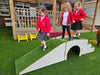 Three young children in red school blazers and dark skirts play on a white wooden climbing tunnel over green artificial turf at an outdoor school play area.