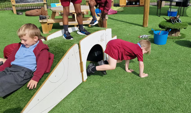 Children playing outdoors on artificial green turf at a playground or daycare, with one child crawling through a white tunnel slide and another sitting on a red bean bag. Toys, tables, and wooden structures are visible in the background.