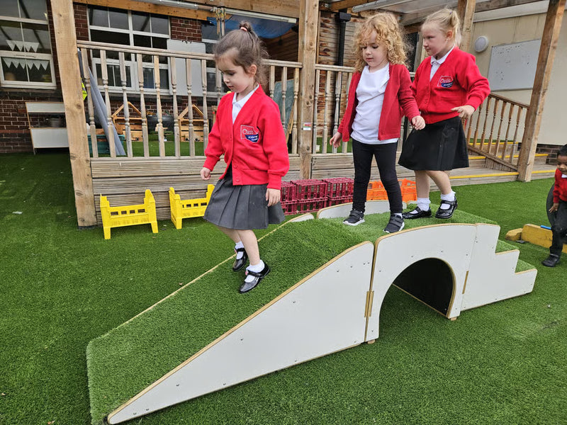 Three young children in red blazers and dark skirts walk across a white wooden tunnel slide on artificial green turf at an outdoor children's play area.