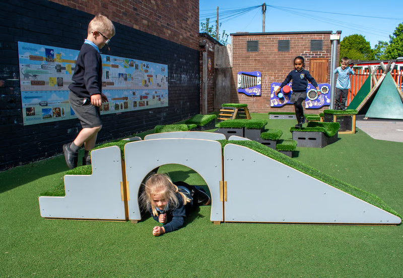 Children playing on an outdoor playground with white wooden climbing structures, artificial green turf, and brick buildings in the background.