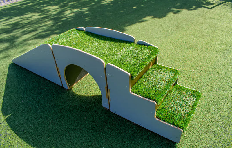 A small outdoor play structure made of white panels with bright green artificial grass on top, including a tunnel opening and two short steps, placed on green artificial turf.