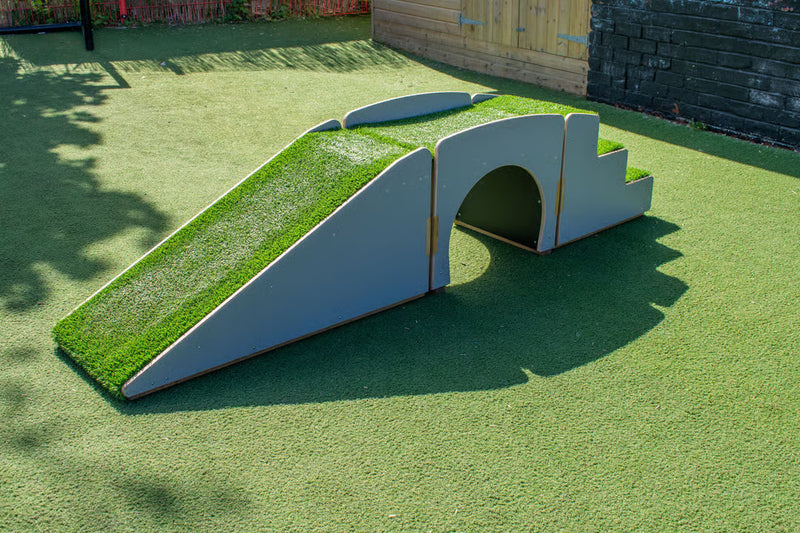 A small outdoor play structure on artificial grass, featuring a grass-covered ramp, a curved tunnel opening, and a small step platform, photographed in a backyard setting.