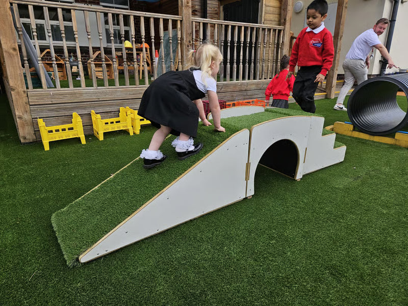 A toddler in a black dress and white shirt climbing a white ramp covered with green artificial grass at an outdoor children's play area. Other children and an adult are visible in the background near other play equipment.