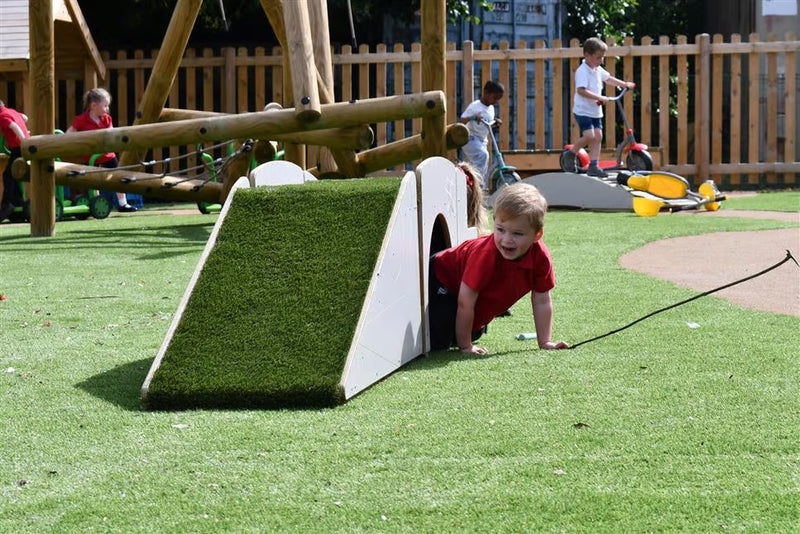 A young child in a red shirt crawls out of a small white play structure with artificial grass on a playground with other children and wooden fencing in the background.
