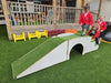 A children's outdoor play structure on artificial grass, featuring a white arched tunnel over a grass-covered incline, with three children in red uniforms playing nearby on a wooden deck.