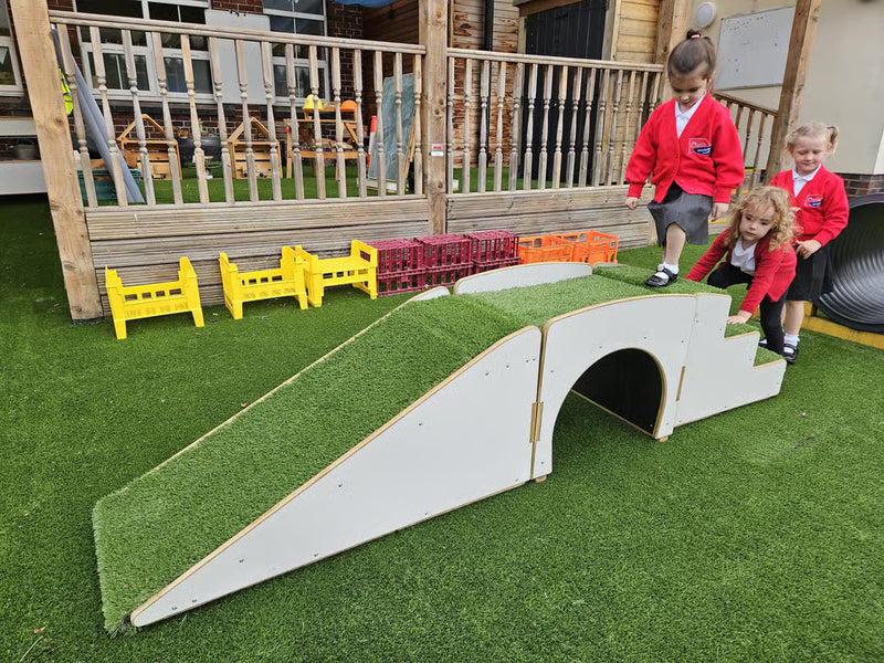 A children's outdoor play structure on artificial grass, featuring a white arched tunnel over a grass-covered incline, with three children in red uniforms playing nearby on a wooden deck.