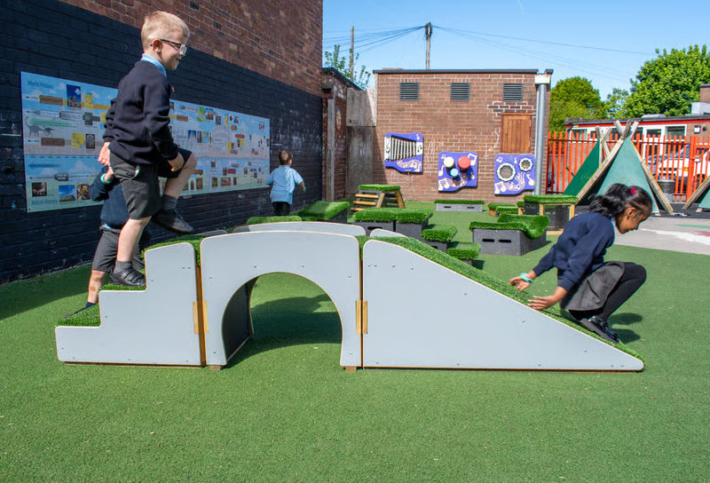 Outdoor playground scene with children using a white wooden climbing bridge and ramp covered in artificial grass, alongside other play equipment and a brick building in the background.
