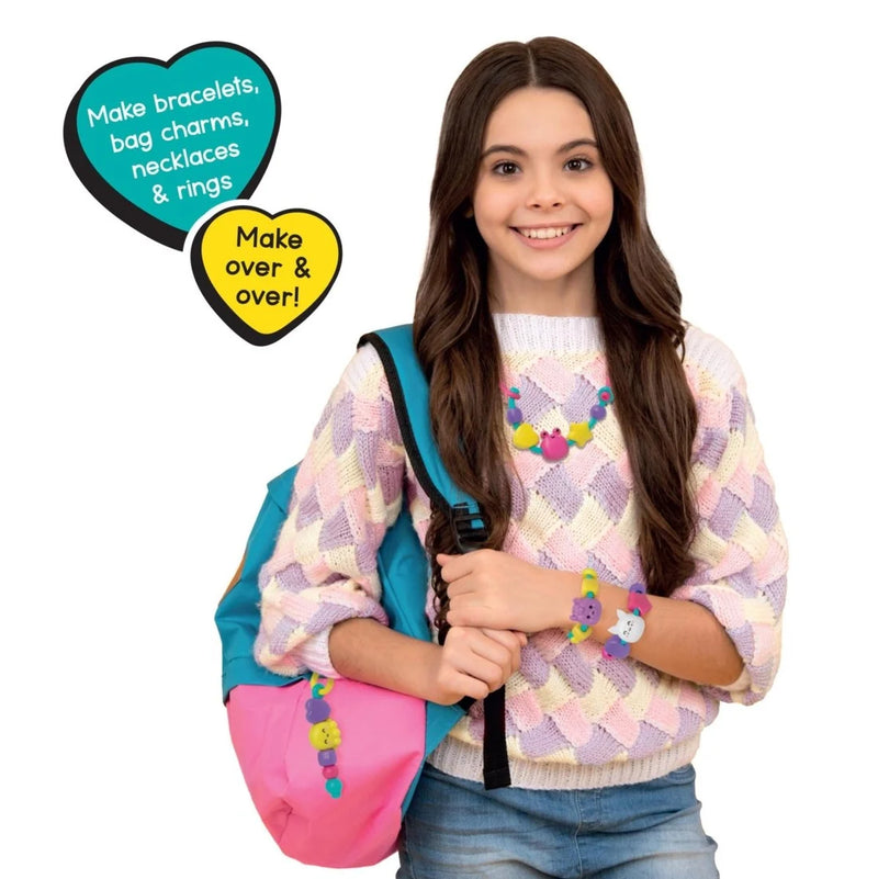 A smiling young woman with long brown hair stands against a white background, wearing a pastel argyle sweater, blue jeans, and colorful craft charms on her bag, necklace, bracelets, and ring. Two heart-shaped callout graphics read 'Make bracelets, bag charms, necklaces & rings' and 'Make over & over!'.