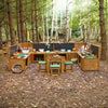 Outdoor wooden camp kitchen set with cooking stations, bowls, bottles, and a small table with green cushioned stools, arranged in a forest clearing with trees and leaf-covered ground.