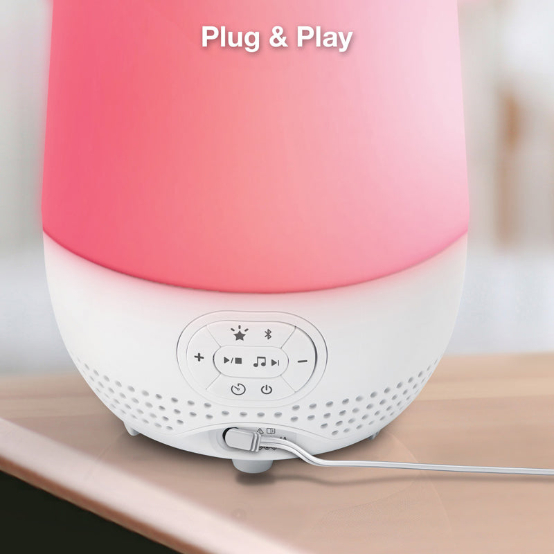 A portable Bluetooth speaker with a pink-to-white gradient upper body and a white control panel base, shown plugged into a wall on a wooden surface with the text 'Plug & Play' at the top.