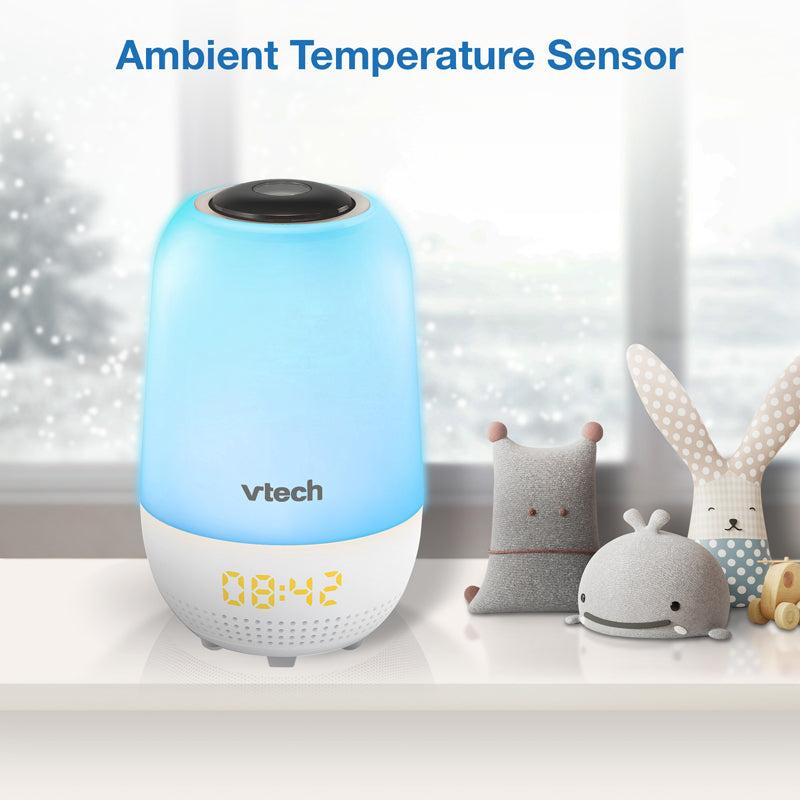 A Vtech ambient temperature sensor alarm clock with a blue and white body, shown on a shelf next to small stuffed toys with a blurred window background. Blue text at the top reads 'Ambient Temperature Sensor'.